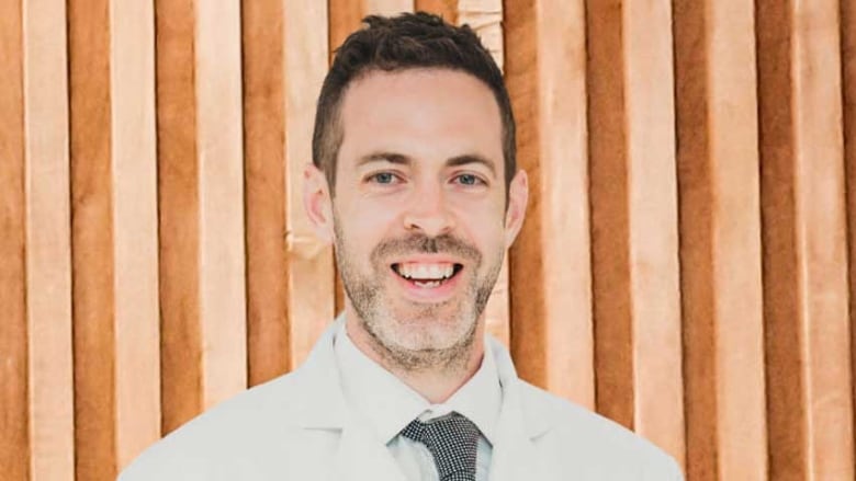 A smiling man with short dark hair and greying beard, earing a collared shirt, tie and a white lab coat.