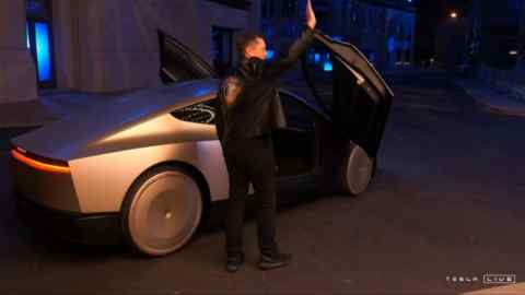 Tesla CEO Elon Musk waving next to his long-promised robotaxi during a launch event at the Warner Brothers studio lot near Los Angeles