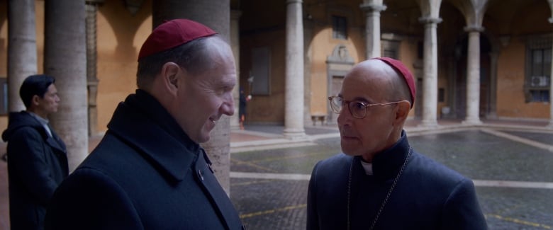 Two men wearing black robes stand in an outdoor square. 