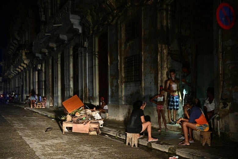 People sit or stand along a road at night during a power outage.