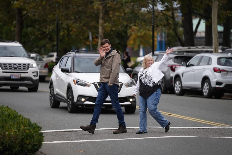 A man and a woman wave as they cross the road.