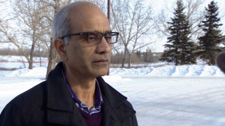 A balding man with glasses wearing a black coat stands outside with snow in the background.