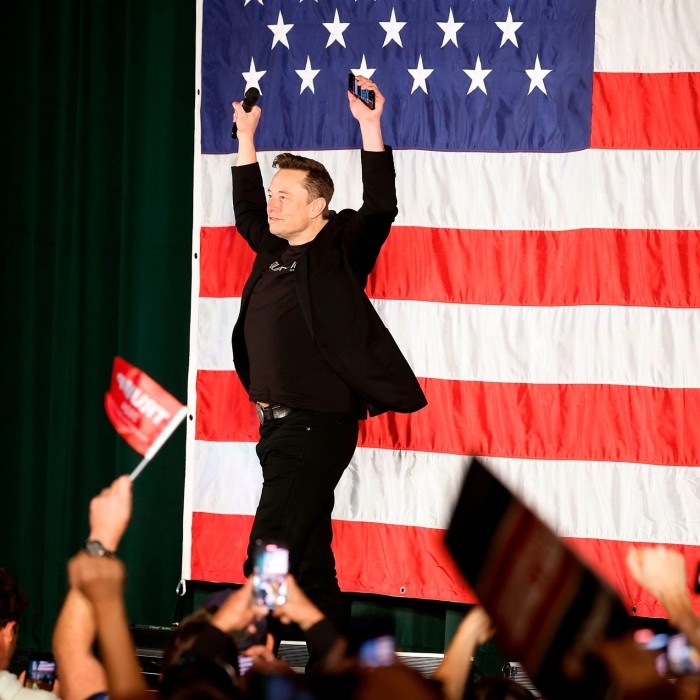 Musk with his arms in the air as he acknowdges the crowds while standing on a stage with an enormous US flag behind him