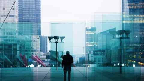 A morning rush hour commuter walks near skyscrapers in the La Defense business district in Paris, France. The person is silhouetted against the backdrop of modern glass buildings and reflections.