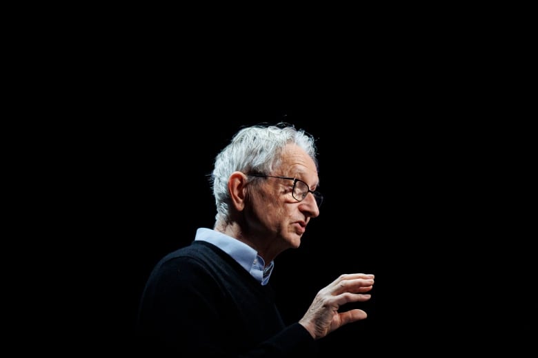 A white-haired man with glasses is seen speaking on stage.