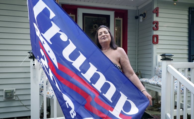 A woman with brown hair, standing in front of a white house, holds a red, white and blue Trump flag.