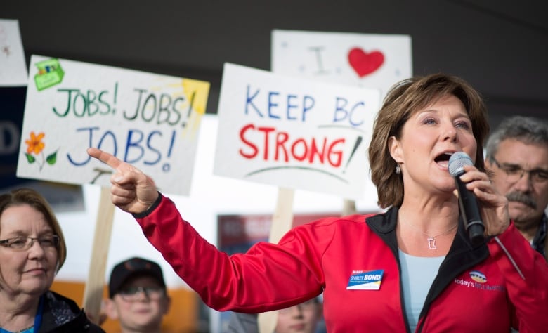 A woman with brown hair and a red jacket speaks into a microphone