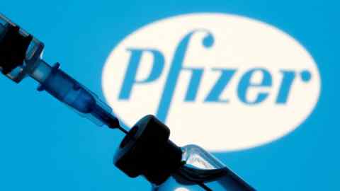 A syringe is inserted into a vial in front of a large, blurred Pfizer logo on a blue background