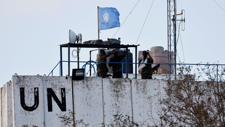 Men in military uniforms stand on a watch tower with a UN flag and the letters "UN" on the side of the building