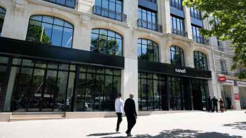 The image shows the Sanofi logo displayed on the facade of the company's headquarters in Paris. The building is modern with large windows and decorative stonework. Two people are walking on the sidewalk, and a group is standing near the entrance.