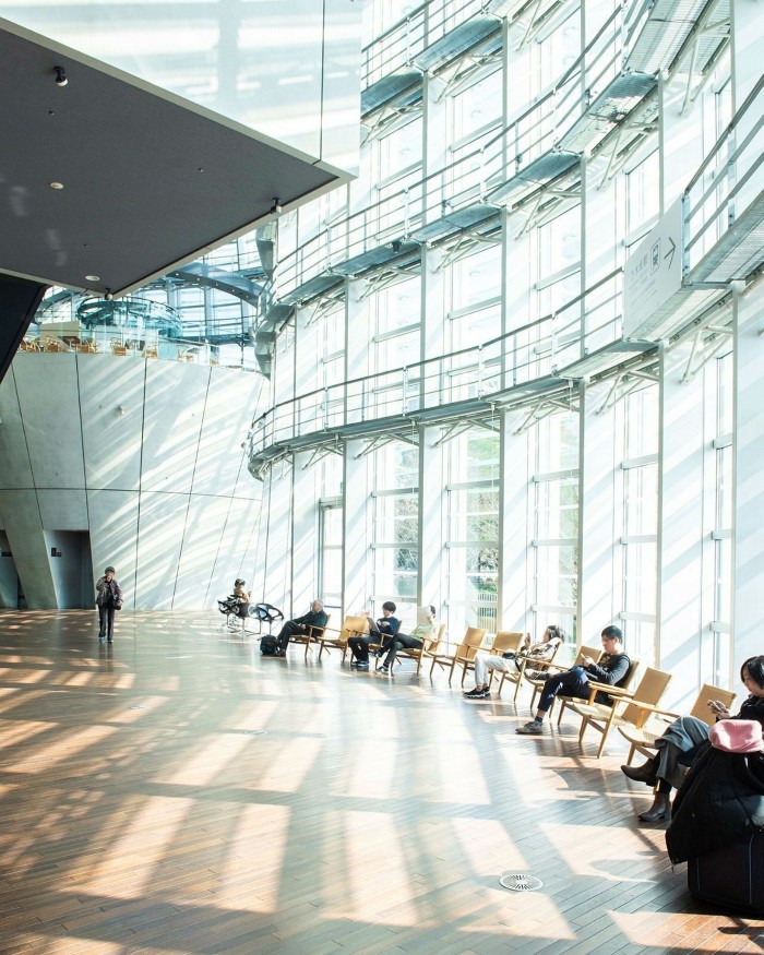 People sitting reading in chairs by a huge curved-glass window inside the National Art Center Tokyo