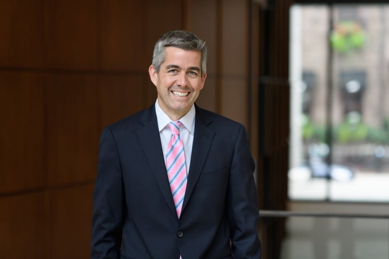 A man wearing a dark navy suit with a pink and blue tie is smiling at the camera. 