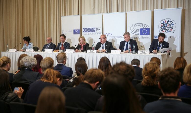 A panel of people sit behind a table onstage as an audiences faces them.