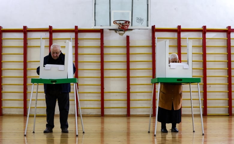 Two elderly people stand behind voting stations inside a gymnasium.