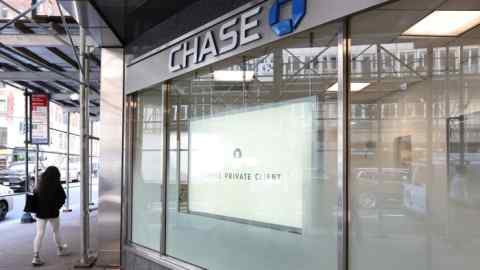 A person walks past a Chase Bank branch in New York City. The bank's signage is prominently displayed, with reflections of cars and pedestrians visible in the glass windows.