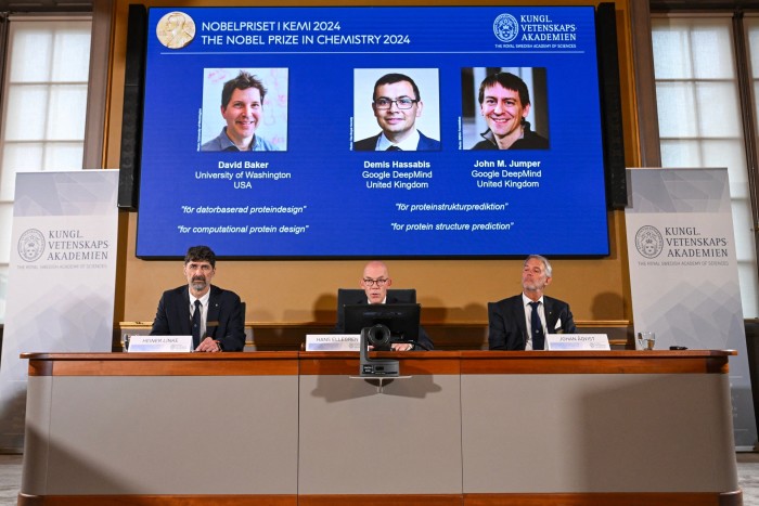 Johan Åqvist, member of the Nobel committee for chemistry; Hans Ellegren, permanent secretary; and Heiner Linke, chairman of the Nobel committee for chemistry, sitting under a screen showing photos of David Baker, Demis Hassabis and John Jumper at the Royal Swedish Academy of Sciences, in Stockholm, Sweden on October 9 2024