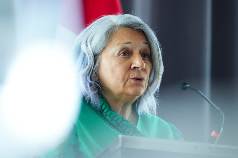 A woman in a green tunic speaks into a microphone. A glare is seen over her right shoulder.