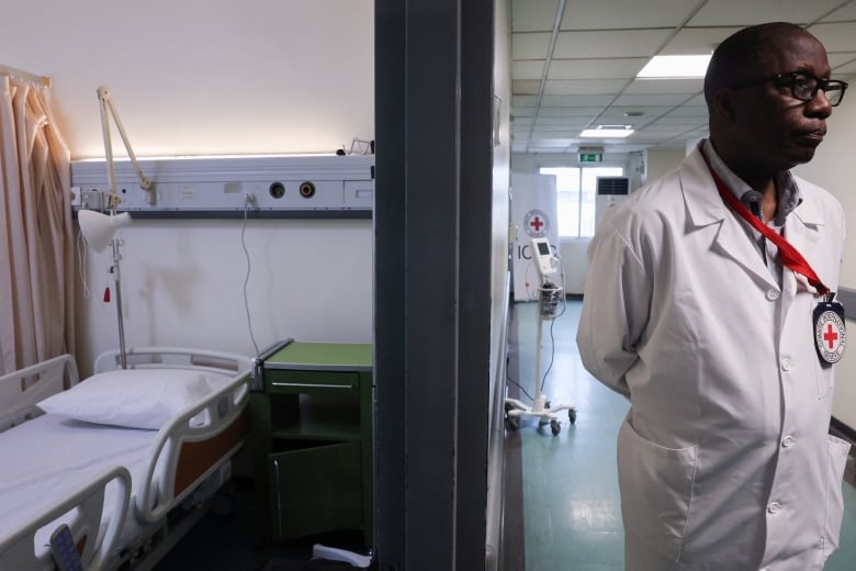 A man in a lab coat with a bright red cross on his badge stands just outside an empty hospital room with his hands behind his back 