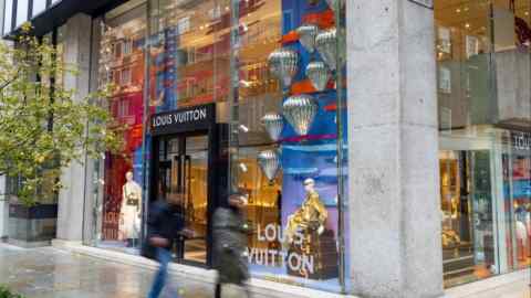 The image shows the exterior of a Louis Vuitton luxury boutique in London. The store has large glass windows displaying mannequins and decorative elements, including metallic balloon figures. Two people are seen walking past the storefront on a rainy day.