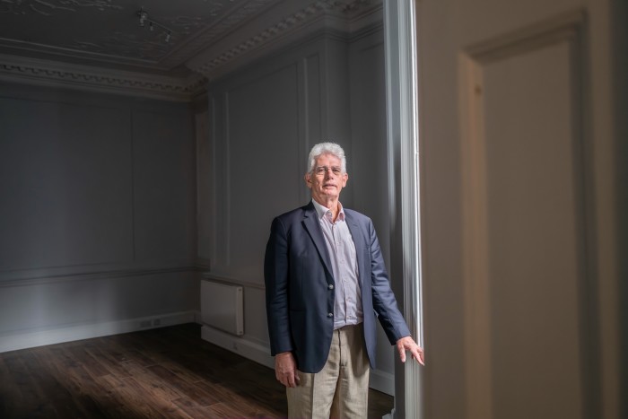 A man with white hair stands near a window in a minimally furnished room, wearing a navy blue blazer and light-colored trousers