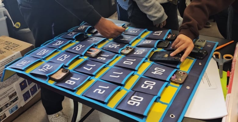 Teen hands are seen slipping cell phones into individual, numbered slots of a 30-pocket holder laying to the side of a teacher's desk.