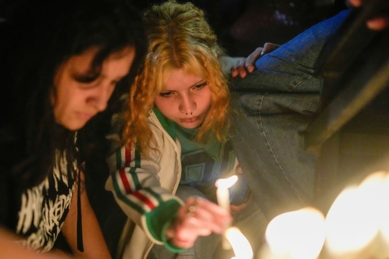 Two young women huddle closely and look mournful as one holds a candle.