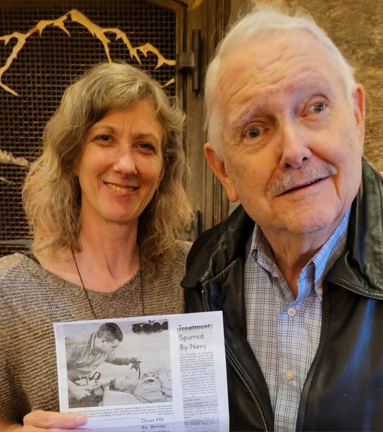 A smiling woman holding a faded newspaper clipping stands next to an older gentleman.  