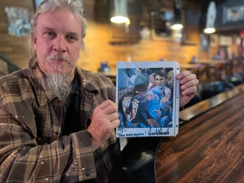 Man in bar holds up picture of a younger version of himself confronting a protester in a Bernie Sanders T-shirt, and the photo is signed in marker by Donald Trump with the message, "TY - YOU ARE GREAT!" 