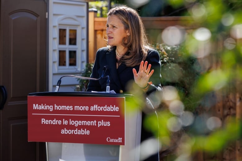 A woman gestures with her hand as she speaks into a microphone at a podium in a suburban backyard.