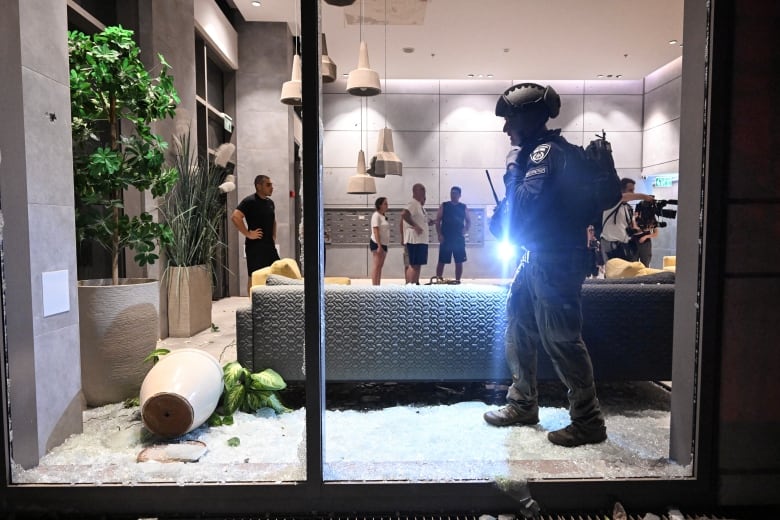 A police officer stands inside a building beside a window that has been shattered. Inside, a plant holder is toppled as people mill about.