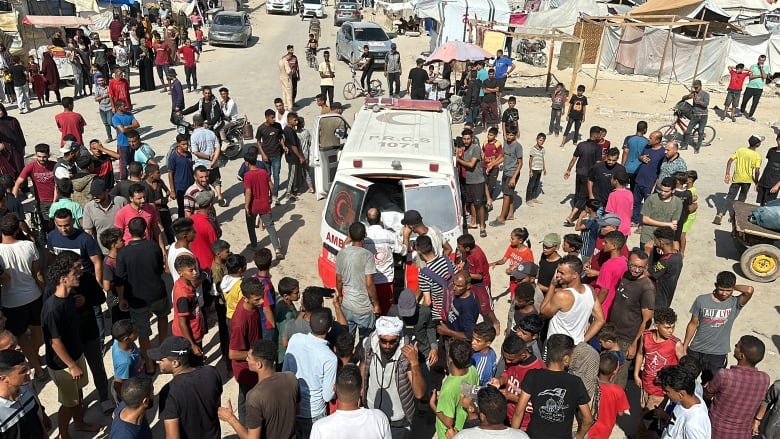 People stand around an ambulance in the middle of a tent camp site.