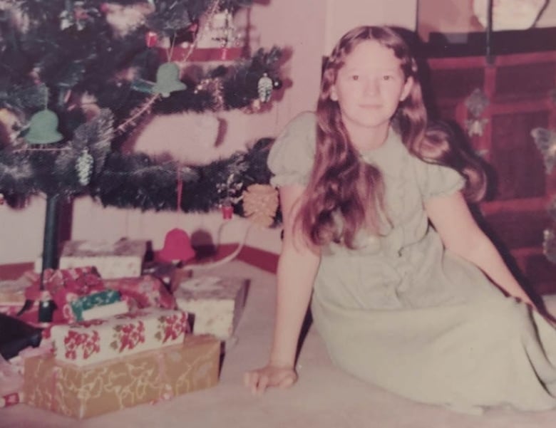 An old looking photo with a 12-year-old girl sitting in front of Christmas tree. 