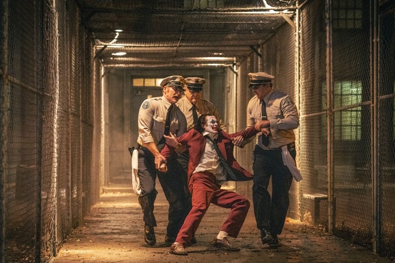 A man in clown makeup struggles as he is forced down a prison hallway by guards. 