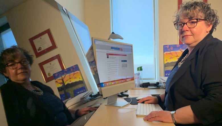 A woman with short curly grey hair, wearing a black blazer and glasses sits in front of one computer monitor but looks over to another on her left.