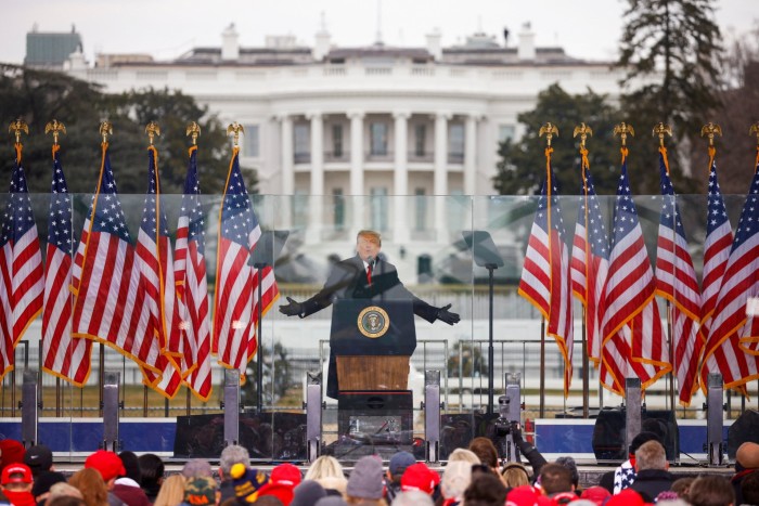 Donald Trump gestures while speaking at a rally in front of the White House. He stands behind a podium with the presidential seal, surrounded by multiple American flags. A crowd of supporters is visible in the foreground.