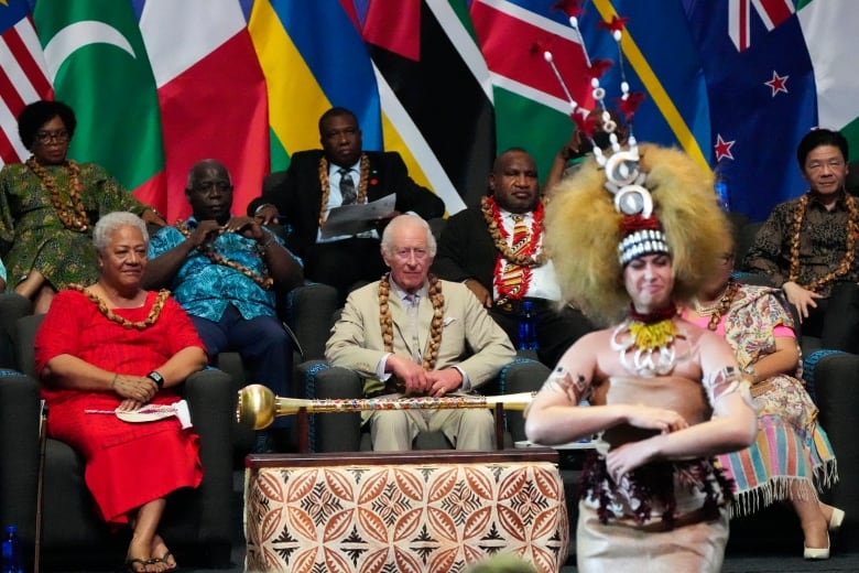 Several people seated in various attire, from suits to dresses, watch an Indigenous dancer who is dressed in costume.