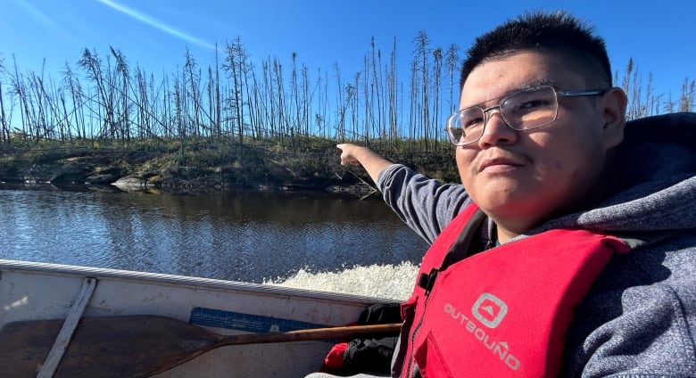 A young man wearing a hoodie, glasses, and a PFD points at a burned section of forest along the bank of a river.