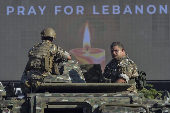 Army soldiers patrol a street in Beirut, Lebanon, riding in a camouflaged military vehicle. Behind them, a large screen displays the message &quot;Pray for Lebanon&quot; with an image of a candle.