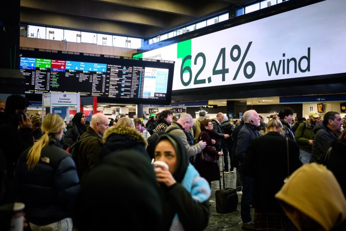 A crowd of travelers awaits under a large digital display showing &quot;62.4% wind&quot; at Euston station in London. 