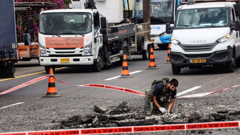 A bearded man in a vest kneels over a section of roadway with large chunks taken out and divots. Behind him, trucks are shown.