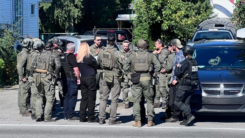 A group of police officers in tactical gear stand in a circle at the side of a road.