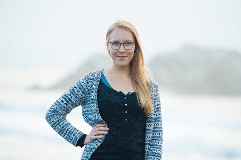 An adult woman with blonde hair and classes poses in front of an outside setting with water and mountains visible in the background.