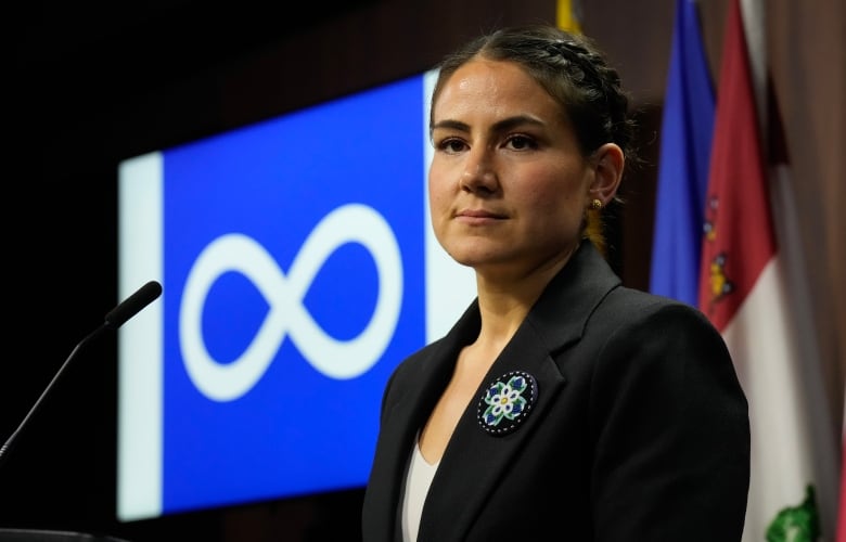 A politician is seen in front of the blue and white Métis Nation flag.