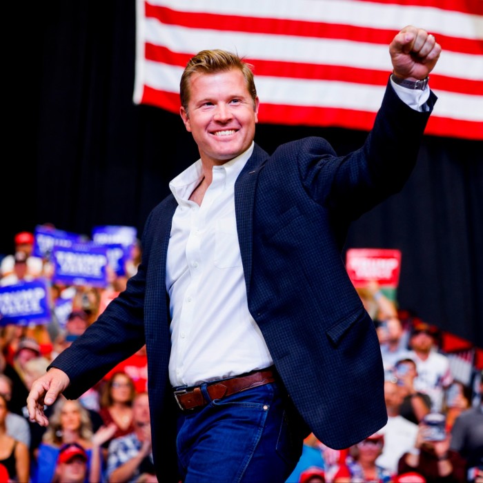 Tim Sheehy walks up to the stage with his fist raised during a rally in Bozeman, Montana, wearing a dark blazer and jeans and with an American flag visible in the background