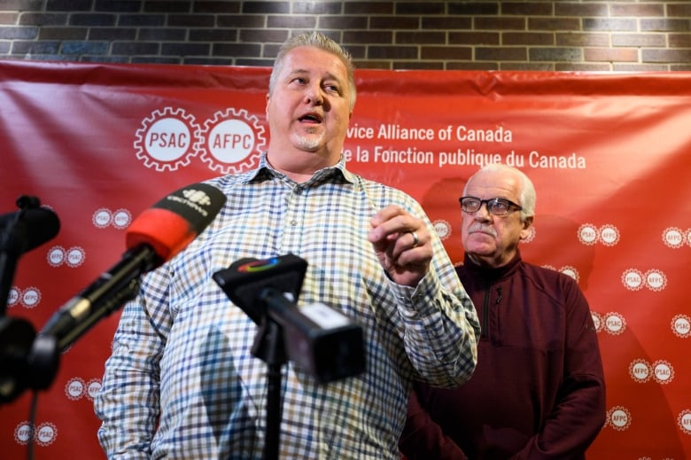 Two union leaders give a news conference. One speaks as the other listens behind him.