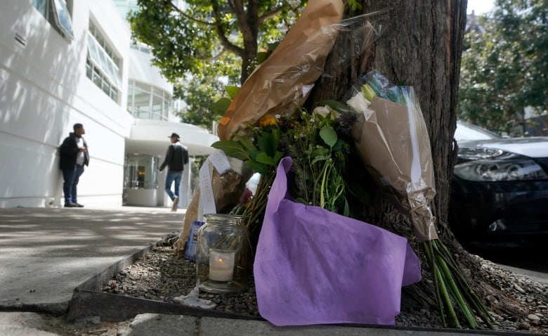 Close-up of a collection of flowers, a candle and purple tissue paper resting against the base of a tree. A white building is visible in the background on the left side, with two people in front of the building. 