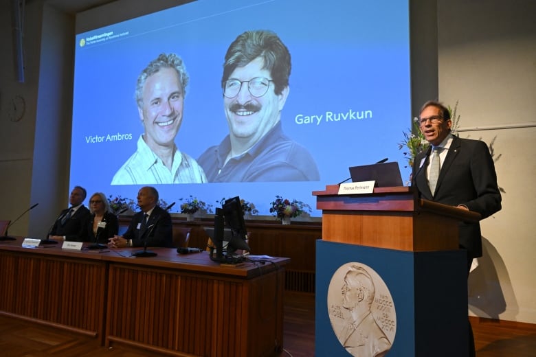 A man stands at a podium, behind him is a screen with the photos of Victor Ambros and Gary Ruvkun.