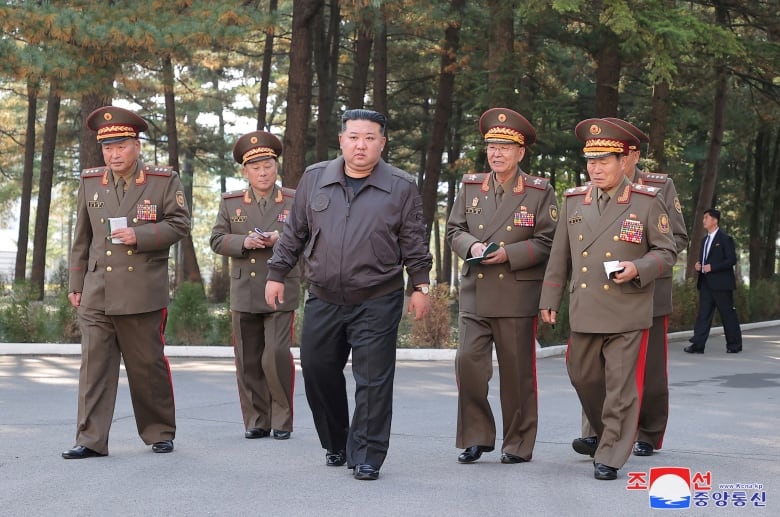 An Asian man in a jacket and dress pants stands between male Asian soldiers in uniform in an outdoor photo.