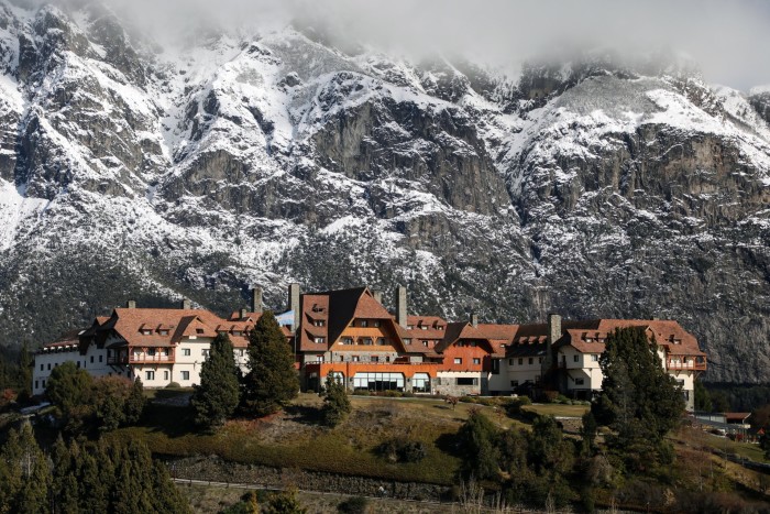 A picturesque lodge with chalet-style architecture, nestled among trees on a mountain slope, framed by dramatic, steep cliffs and snow-capped peaks