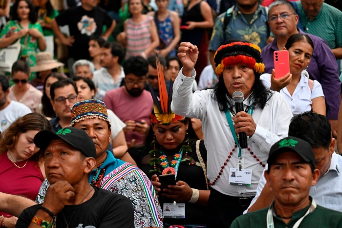 An Indigenous man from the Peruvian Amazon, wearing traditional attire, speaks passionately into a microphone. He is surrounded by a diverse group of attendees, some of whom are also in traditional clothing.